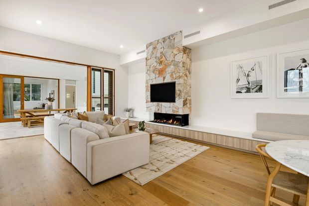 Bright, spacious open-plan living room with warm timber flooring, a large pale sectional sofa, built-in low-profile media cabinetry, and a stone-clad fireplace with integrated television and linear firebox. Timber-framed glazing and doors connect the living area to a dining space, while framed artwork and restrained furnishings create a refined contemporary interior.