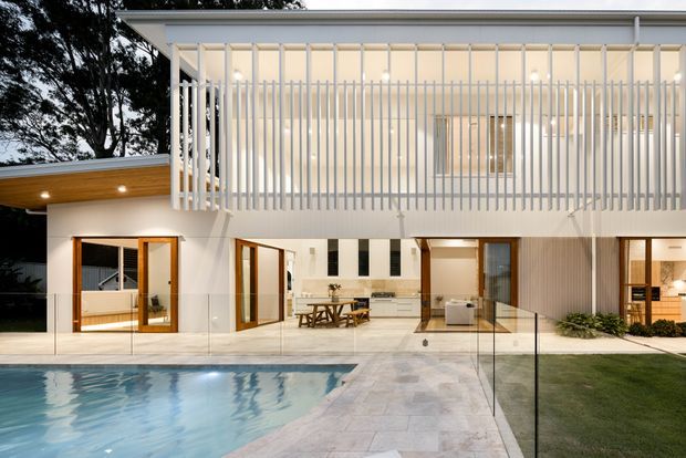 Contemporary two-storey residence viewed from the pool and landscaped yard. The white rendered facade has extensive glazing, warm timber-framed doors, a broad flat roof, and a prominent upper-level vertical slatted screen. A pale stone-paved terrace surrounds the swimming pool, with a sheltered outdoor area and open-plan interior visible through the glazing.