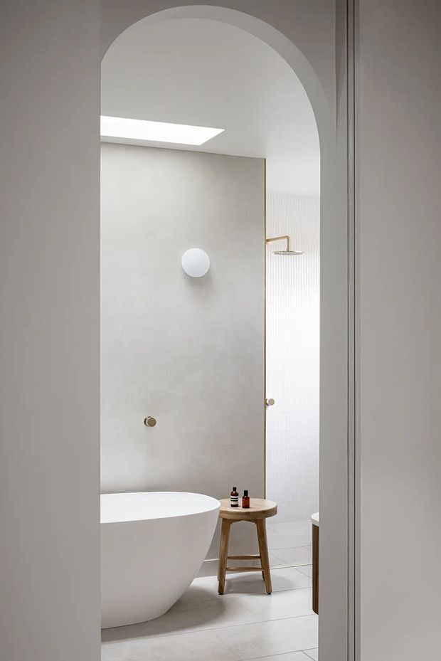 A serene, minimalist bathroom viewed through a tall arched doorway. The space features a freestanding white bathtub, frameless glass shower enclosure with a brass shower fitting, pale textured wall surfaces, white vertical fluted tile, light tiled flooring, and a small natural timber stool with toiletries. A skylight brings soft natural light into the room.