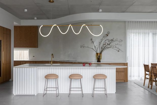 Contemporary open-plan kitchen and dining area with a large white fluted island, three stools, warm timber cabinetry, a stone-look benchtop, concrete-look surfaces, and a sculptural linear pendant light. Sheer curtains and a timber dining setting are visible to the right, while a large decorative branch arrangement adds a natural accent.