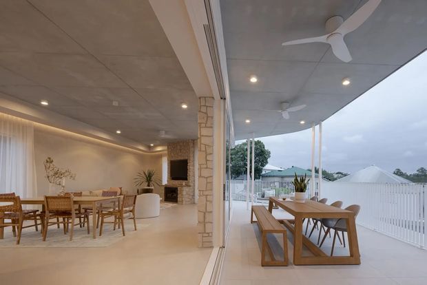 Open-plan indoor-outdoor entertaining area with a dining room and living space opening onto a covered terrace. The interior features a neutral palette, polished light flooring, a stone-clad fireplace wall, recessed lighting, and timber dining furniture. The terrace has a timber dining table, white balustrade, ceiling fans, and views toward neighbouring rooftops.