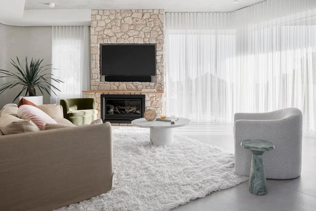 Bright, refined living room with a large cream sectional sofa, sculptural light-grey armchair, shag rug, round coffee table, and a stone-clad fireplace wall with a wall-mounted television. Floor-to-ceiling sheer curtains create an airy setting, complemented by a potted plant and subtle neutral decor.