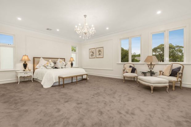 A spacious, elegant bedroom with a large upholstered bed, white bedding, ornate bedside tables and lamps, a chandelier, framed artwork, classic armchairs, and a matching ottoman. The room has soft neutral tones, wall panelling, decorative cornices, large windows, and plush taupe carpeting.