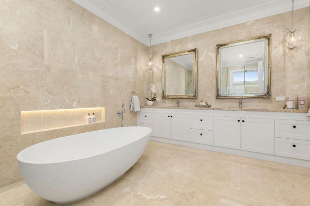 Luxurious, spacious bathroom featuring a freestanding white oval bathtub, extensive beige stone wall and floor finishes, a long white vanity with multiple drawers and twin basins, ornate gold-framed mirrors, wall sconces, recessed lighting, and decorative ceiling cornices.