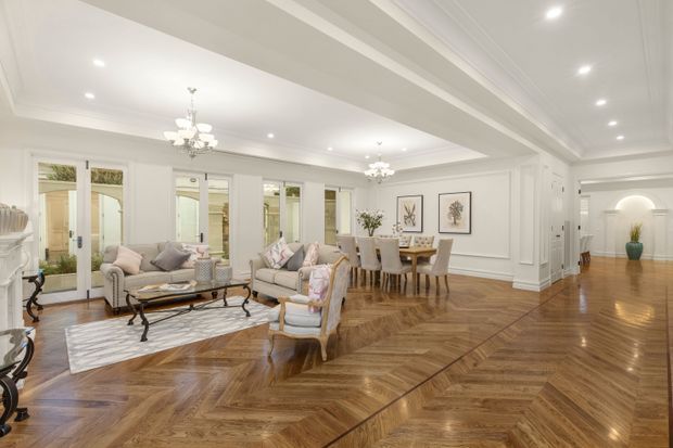 A bright, spacious open-plan formal living and dining area with ornate white wall panelling, layered coffered ceiling details, recessed lighting, chandeliers, tall glazed doors, and polished warm-toned herringbone timber flooring. The room is furnished with upholstered sofas, armchairs, a coffee table, and a formal dining table, creating a refined traditional interior.