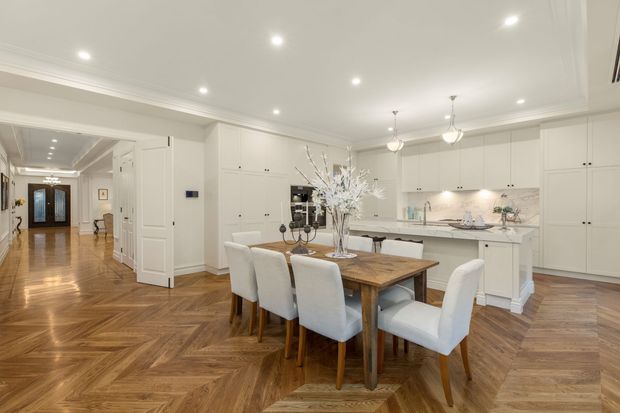 Bright, refined open-plan kitchen and dining area with extensive white built-in cabinetry, a pale stone island and splashback, and a timber dining table surrounded by upholstered chairs. The room features a warm herringbone timber floor, coffered ceiling detailing, recessed downlights, pendant lights, and a hallway leading to a decorative entry door.