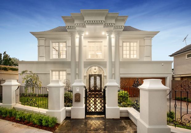Front exterior view of a grand two-storey residence with a symmetrical cream-rendered facade, prominent classical columns, ornate entablature and cornice detailing, arched entry, hipped tiled roof, wrought-iron gate and perimeter fencing. The formal frontage includes paved access and neatly planted garden beds.