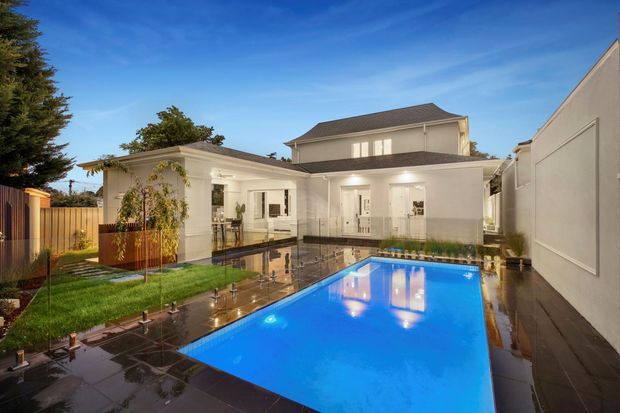 A contemporary two-storey residential home viewed from a landscaped rear courtyard, with a bright rectangular swimming pool, dark stone paving, white rendered walls, a covered outdoor entertaining area, and glazed doors opening to the house. Warm architectural lighting reflects across the wet paving and pool surface at dusk.