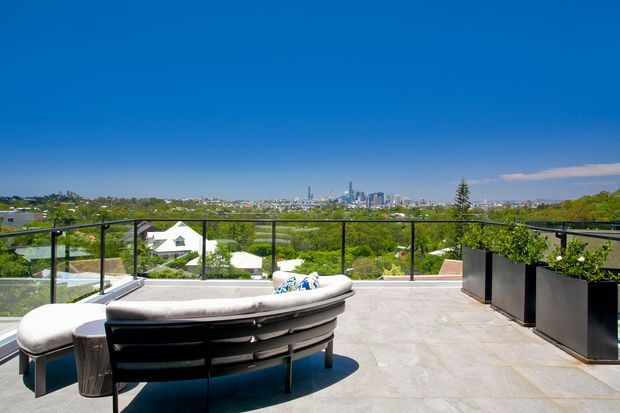 A spacious rooftop or elevated terrace with pale stone paving, black-framed glass balustrades, large dark planters, and a curved outdoor sectional sofa. The terrace overlooks a leafy suburban landscape with a distant urban skyline under a clear blue sky.