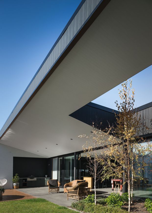 Contemporary residential courtyard viewed from outdoors, featuring a dramatic cantilevered flat roof with a broad white soffit, dark metal-clad walls, extensive floor-to-ceiling glazing, and a paved patio furnished with wicker lounge chairs. A built-in outdoor kitchen or barbecue sits against the dark wall, with landscaped planting and lawn in the foreground.