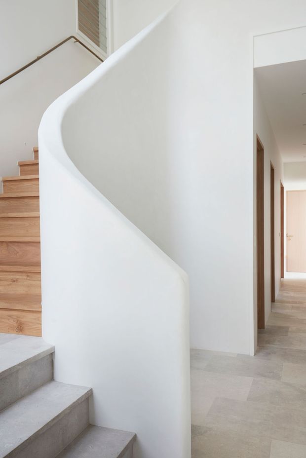 A bright, minimalist interior focused on a sculptural curved staircase with a smooth white plaster balustrade. The stair treads combine warm timber and pale concrete, while a light stone or tile floor extends into a clean hallway with simple timber-framed doorways.
