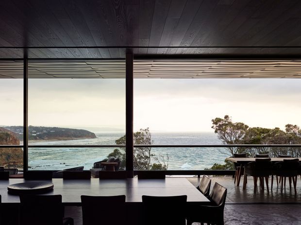 A dark, refined open-plan interior with a large dining table and chairs facing expansive floor-to-ceiling glazing. The room overlooks a rugged coastal inlet and ocean, with a timber-paneled ceiling, dark structural posts and frames, a broad dining area, and a secondary lounge seating arrangement near the windows. The atmosphere is moody and dramatic, with a strong indoor–outdoor connection.