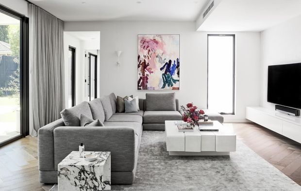 Bright contemporary living room with a large grey sectional sofa, white modular coffee table, marble side table, abstract artwork, floor-to-ceiling glazing with sheer curtains, timber flooring, and a wall-mounted television.