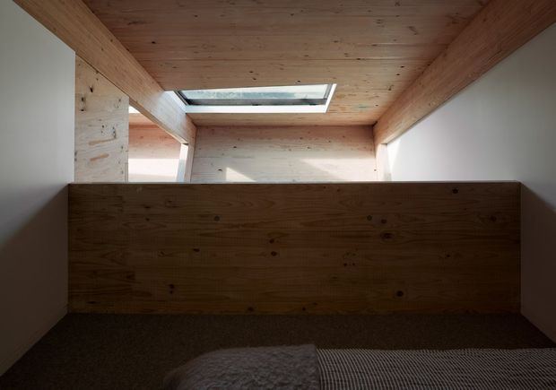 Compact attic-like sleeping space with a low timber partition at the foot of the bed, extensive exposed timber lining across the ceiling and upper walls, white side walls, dark carpet, and a rectangular skylight. The image emphasizes warm natural wood construction and built-in spatial enclosure.