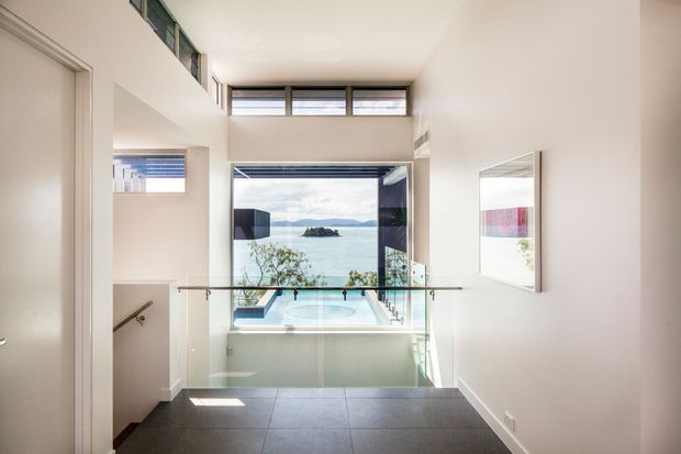 Bright contemporary upper-level landing or hallway overlooking a lower-level room with a spa bath and expansive coastal water views. The space has white walls, dark tiled flooring, high-level clerestory windows, a large opening to the view, a frameless glass balustrade, and a wall mirror. A stair descends on the left.