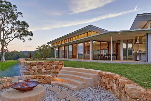 A contemporary single-storey residence set into a landscaped site, viewed from the garden. The home has broad floor-to-ceiling glazing, low-pitched projecting roof forms with clerestory-like upper glazing, warm timber-toned framing, and an open-plan interior visible through the glass. The foreground features a stone retaining wall, curved concrete steps, gravel paving, and a built-in circular fire bowl.