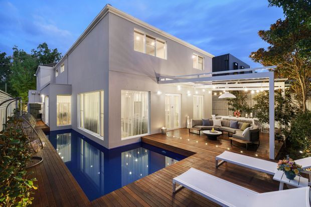 A contemporary multi-level home viewed from a timber deck at dusk, centred on a reflective rectangular swimming pool and an outdoor entertaining area. The pale rendered facade has large glazed doors and windows, while a white pergola with integrated lighting shelters an outdoor lounge. Timber decking wraps around the pool and extends to a dining area, creating a polished indoor-outdoor connection.