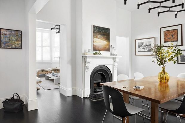 Bright, refined dining room in a traditional-style home, featuring a large timber dining table, contemporary black chairs, white walls and trim, dark flooring, framed artwork, and an ornate white mantel with a black fireplace. A living room is visible through the open doorway, and sculptural black track lighting hangs overhead.