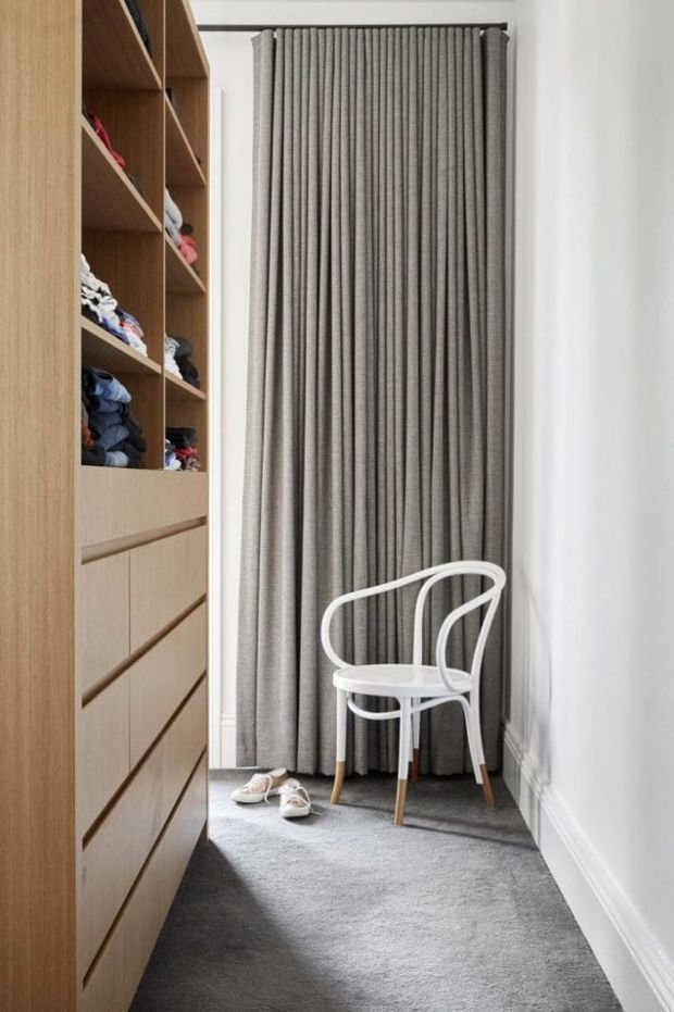 A narrow walk-in wardrobe or dressing area with floor-to-ceiling built-in timber shelving and drawers along the left wall. Grey full-height pleated curtains cover the rear opening, with a white bentwood-style chair and shoes on a soft grey carpet. White walls, ceiling, and skirting create a clean, understated interior.