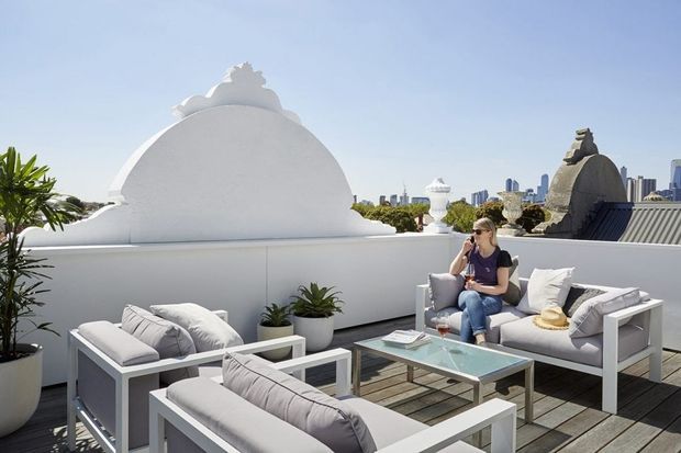 Bright rooftop terrace with white outdoor lounge furniture arranged around a glass-topped coffee table. The terrace has light timber decking, white rendered parapet walls, sculptural decorative architectural elements, potted plants, and views toward a distant urban skyline. A seated person is present, but the space and outdoor furnishings are the main subject.