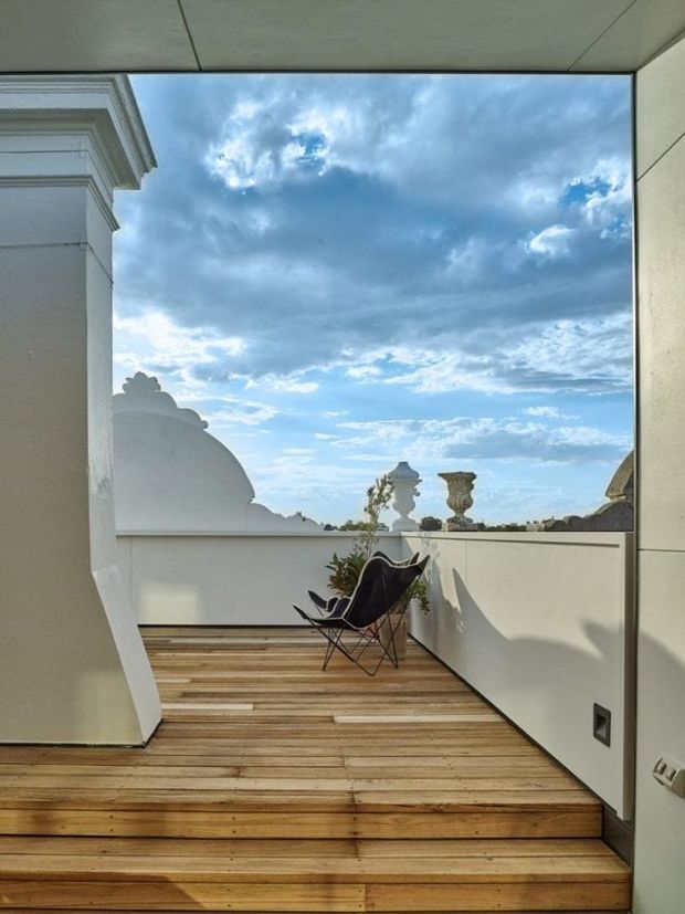 A compact elevated terrace or balcony viewed from an adjoining interior, featuring stepped timber decking, white rendered walls, a low parapet, sculptural classical rooftop elements in the distance, and two black sling chairs with a potted plant. Large open glazing frames a dramatic blue and cloudy sky, creating a bright architectural outdoor setting.