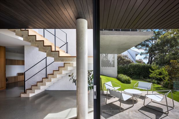 A contemporary interior opening onto a landscaped courtyard. The main view features a sculptural open-riser staircase with pale concrete treads, timber edging, slim black metal balustrades, a white textured wall, polished grey concrete flooring, a dark timber-lined ceiling, and a prominent cylindrical concrete column. Through the full-height glazed opening is a paved outdoor sitting area with modern white chairs and a small table, lawn, planting, and a cantilevered concrete structure.
