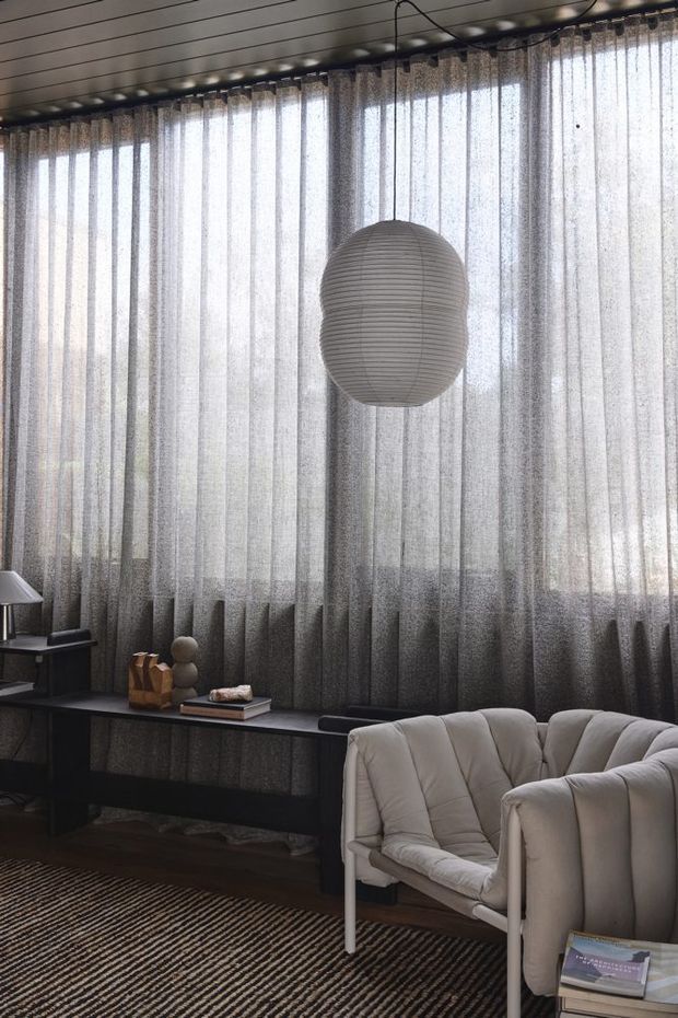A moody contemporary living room with floor-to-ceiling sheer textured curtains, a dark built-in or console-style ledge, sculptural decor, and a large spherical pendant light. A light upholstered lounge chair and sofa sit over a dark striped rug, with a linear timber-toned ceiling adding texture.