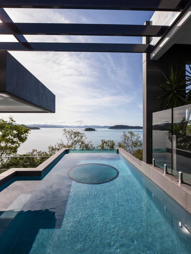 A contemporary elevated outdoor swimming pool overlooking a broad coastal bay with islands. The pool is framed by dark structural beams and concrete walls, with glass balustrading, tiled or rendered pool surrounds, and an integrated circular spa. The scene has expansive water views and a bright open sky.