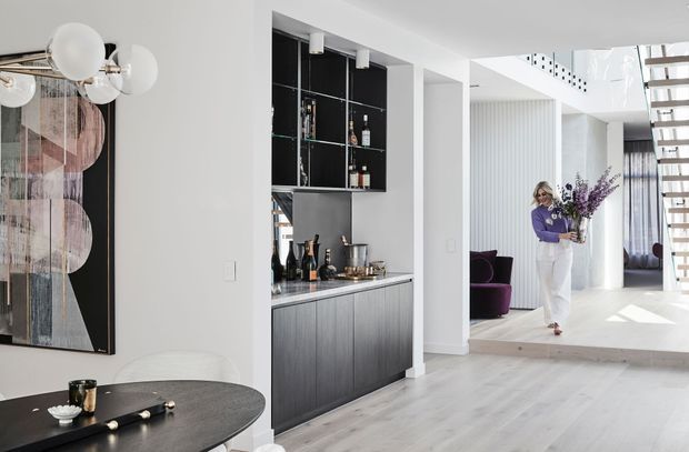 A bright, contemporary open-plan interior showing a dining area in the foreground, a built-in bar and drinks cabinet at centre, and a split-level living area with a floating staircase in the background. The space features white walls, pale timber flooring, dark cabinetry, sculptural pendant lighting, and abstract wall art.