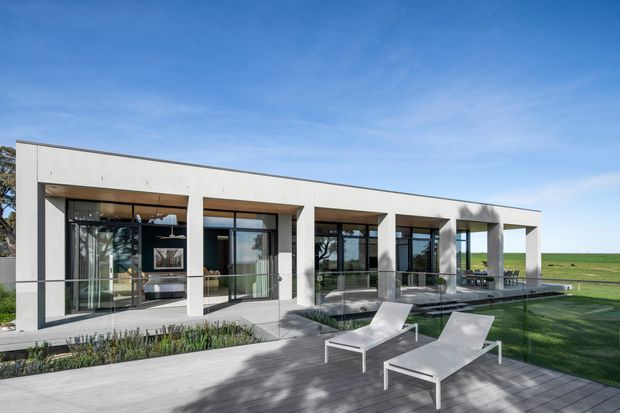 A low, rectilinear modern house viewed from the outdoor deck, with a flat roof, pale rendered facade, deep recessed openings, extensive dark-framed glazing, and glass balustrades. White sun loungers sit on a timber-toned deck beside a landscaped lawn and open rural field. The image is bright and minimalist, emphasizing the building exterior and outdoor entertaining area.