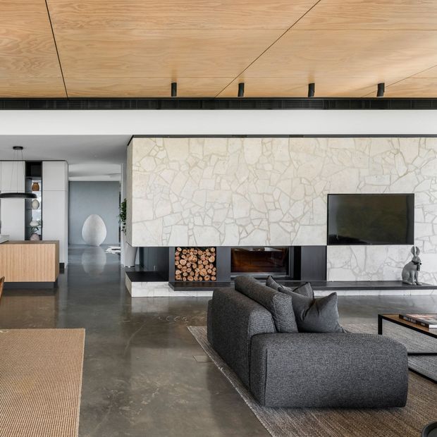 A refined open-plan living room with a large dark grey sectional sofa facing a television and fireplace integrated into a pale irregular stone feature wall. The room has a polished concrete floor, warm timber ceiling with linear joints and concealed ventilation, dark built-in cabinetry, a partially visible kitchen, and minimalist furnishings. The palette is neutral with warm timber accents and strong textural contrast.