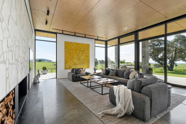 Contemporary living room with expansive floor-to-ceiling glazing overlooking a rural landscape. The room features a grey sectional sofa arrangement, low black-framed coffee tables, a large neutral rug, a white feature wall with a yellow artwork, a sculptural stone fireplace wall with exposed firewood, and a warm plywood-lined ceiling. Polished concrete flooring and slim black window frames create a refined modern interior with strong indoor-outdoor connection.