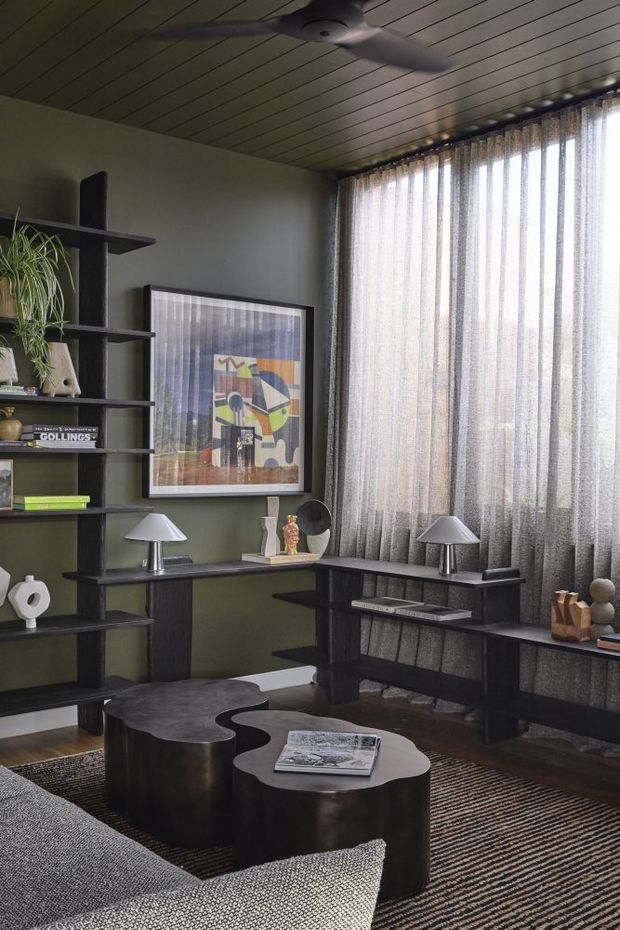 A dark, refined living room with olive-green walls, extensive black built-in open shelving and a long integrated desk or console beneath full-height sheer curtains. A sculptural dark coffee table, grey upholstered sofa, table lamps, framed abstract artwork, books and decorative objects furnish the room. The ceiling has narrow timber-look linear panelling and a contemporary ceiling fan, while a striped rug anchors the seating area.