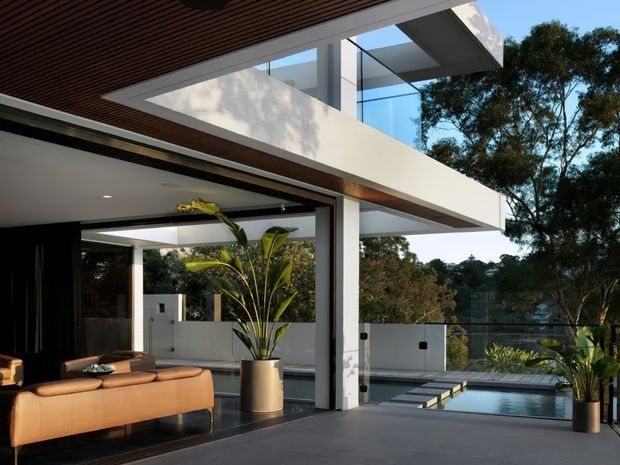 Contemporary multi-level residence with strong indoor–outdoor connection, shown from a covered terrace or living area. The image features a warm timber-slat soffit, crisp white rendered and concrete-like volumes, extensive floor-to-ceiling glazing, dark flooring, a tan leather sofa, and a landscaped pool terrace beyond glass balustrades. Mature trees surround the property, while a cantilevered upper level creates a sheltered outdoor living space.