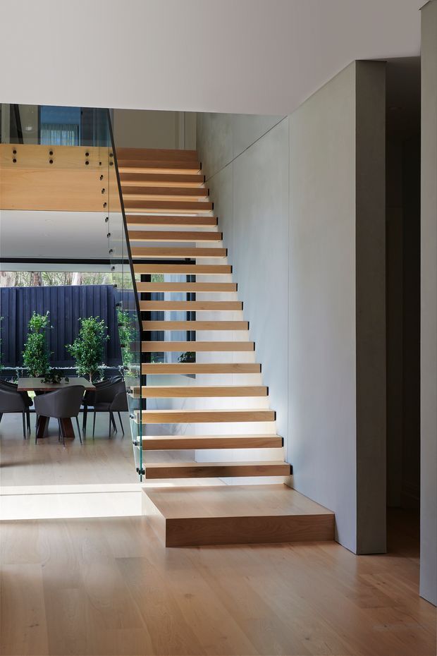 Contemporary interior view centred on a floating timber staircase with clear glass balustrade and slim dark handrail. The staircase rises beside a smooth light wall and connects to an open dining area visible behind it, with timber flooring, large glazing, and a dark exterior fence beyond. Clean lines, concealed structure, and natural timber create a refined, minimalist atmosphere.