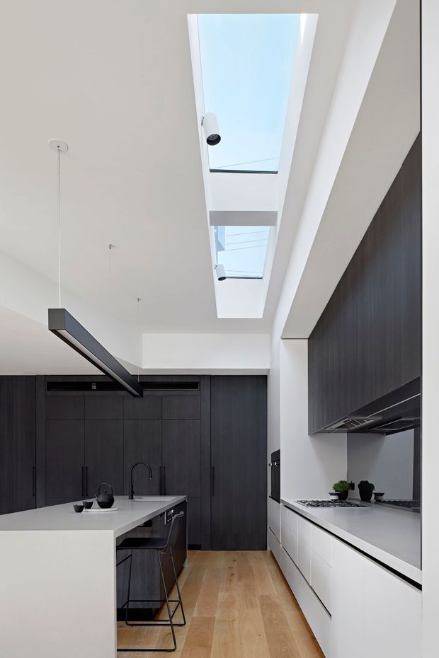 Contemporary galley kitchen with a large white waterfall-edge island, dark charcoal timber cabinetry, white lower cabinets and benchtops, integrated appliances, and a linear black pendant light. Two elongated skylights bring abundant natural light into the otherwise restrained interior, while light timber flooring adds warmth.