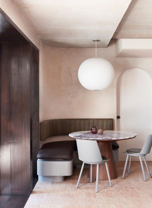 A compact, refined dining nook with a built-in curved banquette upholstered in dark brown leather, a round marble-topped table, sculptural white dining chairs, and a spherical pendant light. The space features warm pink-beige plaster walls, pale timber ceiling beams, dark timber built-in joinery, and a pale terracotta-toned tiled floor. An arched doorway and softly rounded forms add a contemporary, softly sculpted character.
