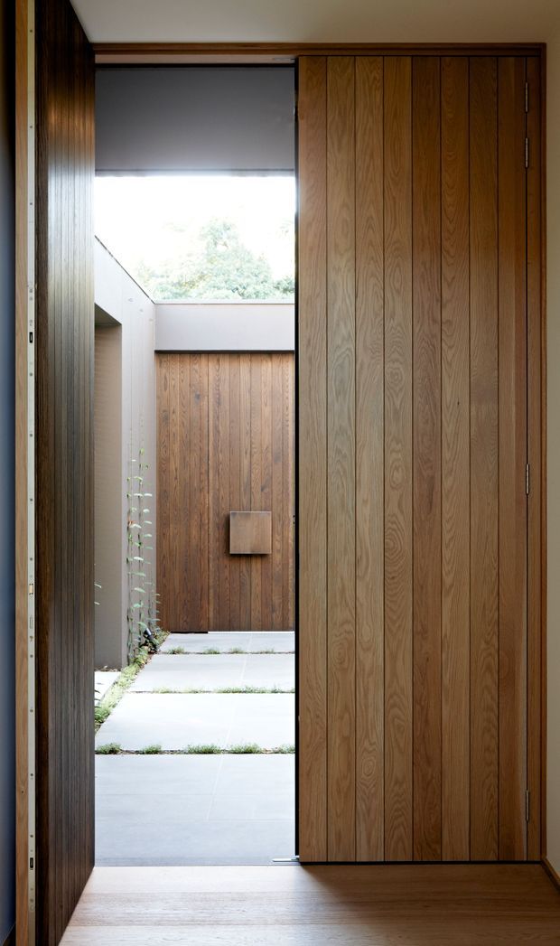 A narrow, refined entryway framed by warm vertical timber wall panelling and a large timber pivot door. The view opens toward a minimalist paved courtyard with additional timber cladding, a high boundary wall, and a partially screened skylit opening. The interior has timber flooring, dark timber trim, and a restrained architectural composition with strong indoor–outdoor connection.