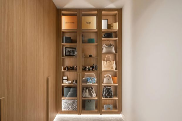 A warmly lit built-in display and storage wall, featuring full-height vertical timber panelling on the left and a timber-framed shelving unit with integrated lighting. The shelves display handbags, shoes, bottles, boxes, framed photographs, and small decorative objects. A softly illuminated floor-level recess and sculptural ceiling detail add a refined, bespoke feel.