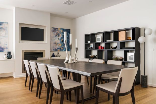A refined open-plan dining and living area dominated by a large dark dining table with upholstered dining chairs. The room includes warm timber flooring, a recessed wall-mounted television above a fireplace, abstract artwork, sculptural table decor, and a large dark built-in shelving unit displaying decorative objects. Neutral white walls and contemporary furnishings create a polished, spacious interior.
