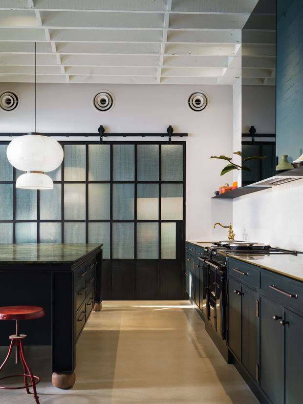 A dramatic industrial-style kitchen with dark blue-black cabinetry, a central island, brass hardware and tapware, and a large black-framed frosted-glass sliding door. The space features a white-painted exposed beam ceiling, circular ceiling vents, white globe pendant lights, polished concrete flooring, and a bold black-and-white palette with warm metallic accents.