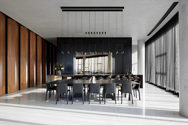 A dramatic, spacious open-plan dining and kitchen interior with a long dark dining table and multiple upholstered chairs in the foreground. A large black kitchen island and dark built-in cabinetry sit against a backdrop of warm vertical timber wall panelling, while full-height sheer curtains and expansive glazing flood the polished light floor with natural light. A linear suspended chandelier hangs above the dining table.