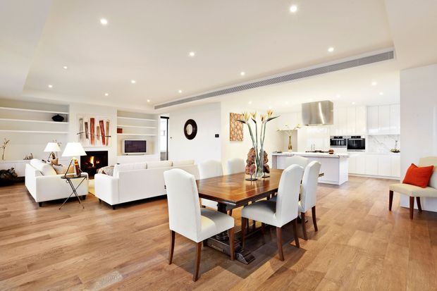 Bright open-plan contemporary interior combining a dining area, living room and modern white kitchen. The space features warm timber flooring, a large timber dining table with upholstered chairs, white sofas, recessed ceiling lighting, built-in shelving, a fireplace and extensive white cabinetry. The room has a clean, spacious and inviting atmosphere.