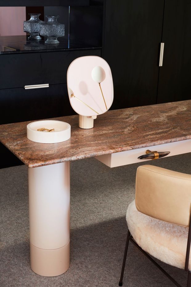 A close-up of a refined home office or dressing desk area. A sculptural stone-look desk with a cylindrical white pedestal leg and a suspended drawer is set against dark built-in cabinetry with slim light handles. The desk holds a rounded tabletop mirror and a small white dish, while a curved upholstered chair sits in the foreground. The palette is dark, warm, and luxurious, with tactile stone, carpet, metal, and soft upholstery.