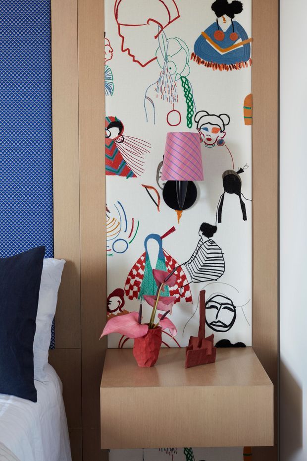 Close-up interior view of a compact bedroom bedside area. A light timber floating bedside cabinet is positioned beside a partially visible bed, with a dark navy cushion and white bedding. Behind it is a tall decorative illustrated wall panel featuring colourful abstract figures and faces. Small sculptural decor and a red vase with pink flowers sit on the cabinet. The room has a playful, artistic, contemporary character with warm timber joinery and bold graphic accents.