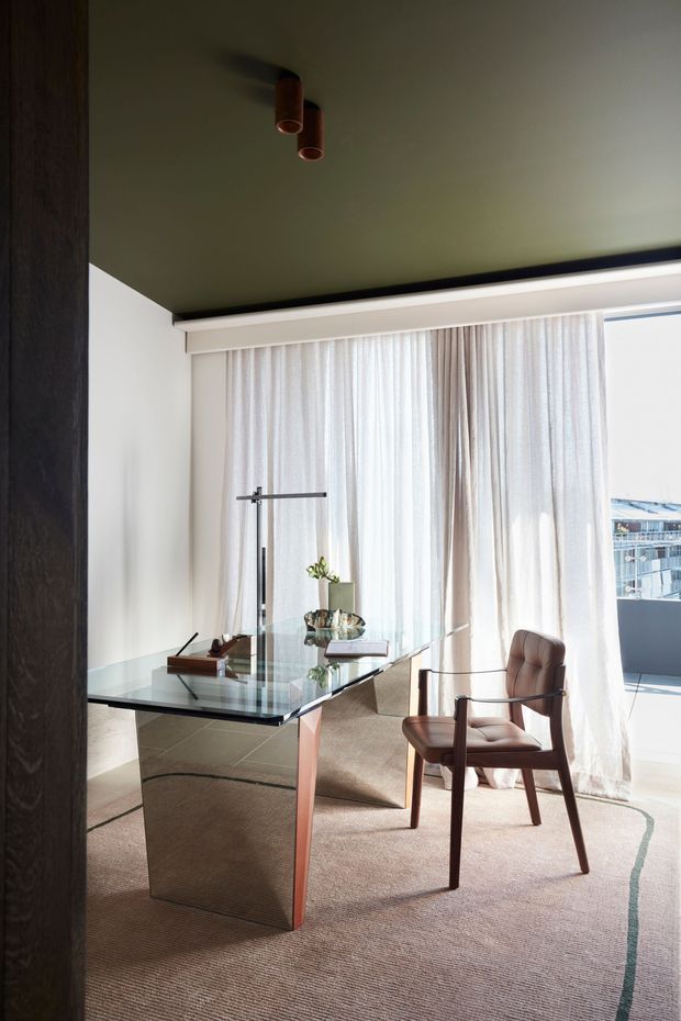 A refined home office with a glass-topped desk featuring a sculptural metallic base, a wooden-framed upholstered armchair, a slim task lamp, and a small plant. Full-height sheer curtains provide soft daylight beside a balcony or terrace, while a dark olive ceiling, white walls, dark timber framing, and a neutral textured rug create a warm, sophisticated atmosphere.