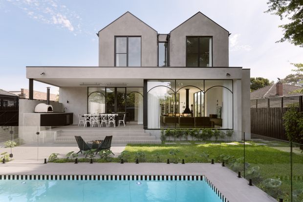 Rear exterior view of a contemporary two-storey residence with two gabled upper volumes, a broad covered terrace, extensive arched glazing, and a white rendered facade. The terrace includes an outdoor kitchen and dining area, opening onto a landscaped lawn and swimming pool enclosed by frameless glass fencing.