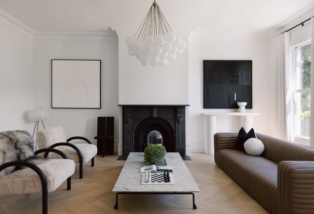 Bright, refined living room with white walls, ornate dark fireplace surround, herringbone timber flooring, sculptural pendant light, contemporary ribbed sofa, boucle armchairs, marble coffee table, and restrained monochrome artwork. Tall windows bring in abundant natural light.