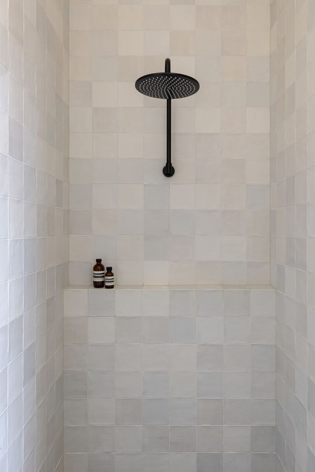 A compact walk-in shower enclosure lined floor-to-ceiling with pale square tiles, including a tiled half-height ledge at the rear. A large round matte-black rainfall showerhead and matching vertical riser provide a strong contrast, with two small amber bottles placed on the ledge. The space is clean, minimal, and softly lit.
