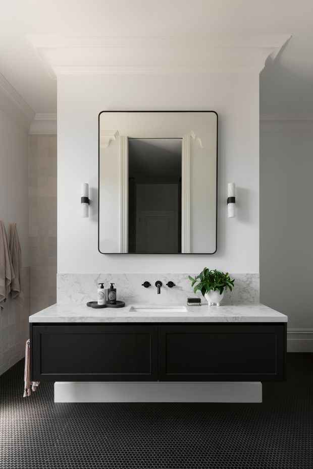 A refined bathroom vanity installation with a floating black double-width cabinet, white marble countertop and integrated basin, black wall-mounted tapware, and a large rounded-corner mirror with a dark frame. The room features white walls with decorative cornice detailing, traditional architectural trim around the reflected doorway, black-and-white mosaic tile flooring, wall sconces, and a small potted plant.